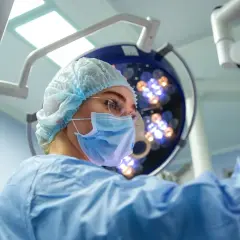 Chirurg w masce i czepku na tle lampy operacyjnej.